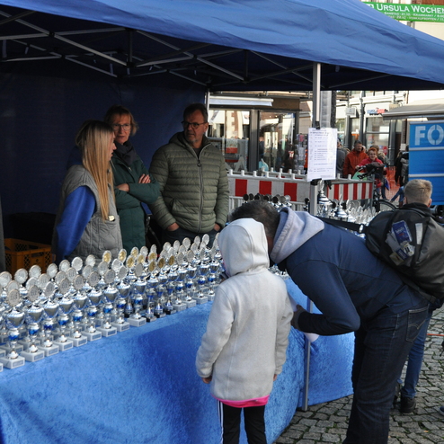 Menschen betrachten Pokale an einem Stand unter einem Zeltdach auf einem Markt.