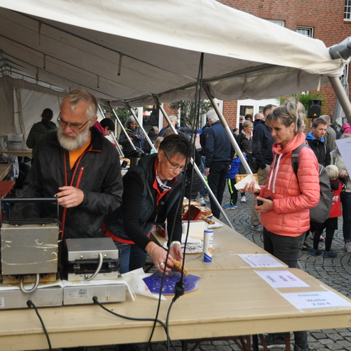 Zwei Personen bereiten Essen an einem Tisch unter einem Zelt, während Besucher im Hintergrund warten.