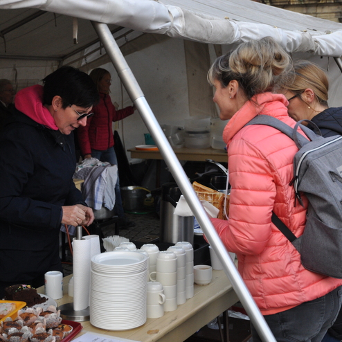 Frauen stehen an einem Tisch mit Getränken und Snacks unter einem Zelt auf einem Markt. Eine Frau bedient eine Kundin.