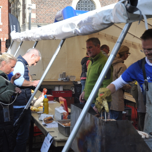 Menschen stehen an einem Verkaufsstand mit Bratwürsten und Getränken unter einem Zelt in einer belebten Umgebung.