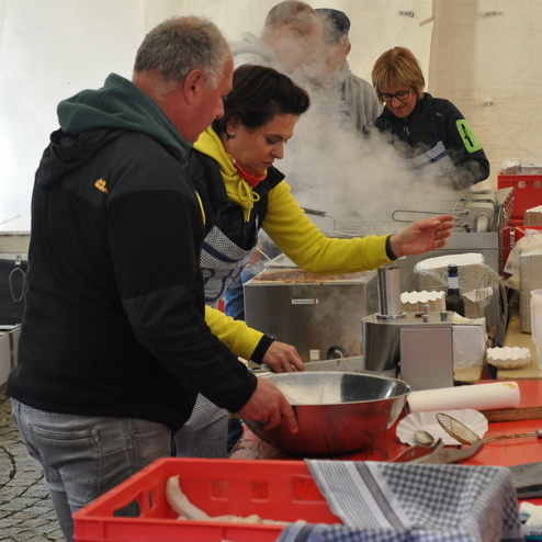 Vier Personen bereiten in einem Zelt dampfendes Essen vor, umgeben von Küchengeräten und Zutaten auf Tischen.
