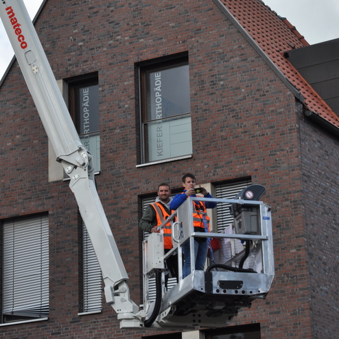 Arbeiter in einem Hochlift vor einem Backsteingebäude, beobachten die Umgebung und halten eine Kamera.