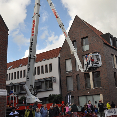 Bauarbeiter in einer Hebebühne reparieren ein Gebäude, während Zuschauer hinter einer Absperrung beobachten.