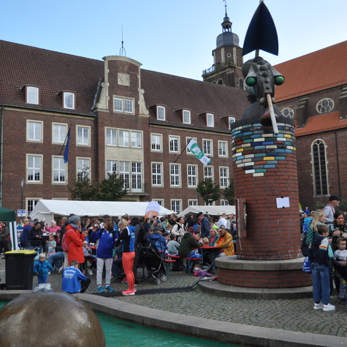 Menschenmenge vor einem alten Geb&auml;ude mit einer skulpturalen Figur und St&auml;nden auf einer Veranstaltung im Freien.