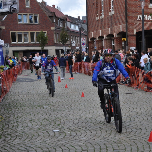 Zwei Radfahrer passieren eine mit Pylonen markierte Strecke, umgeben von Zuschauern und einer festlichen Atmosph&auml;re.