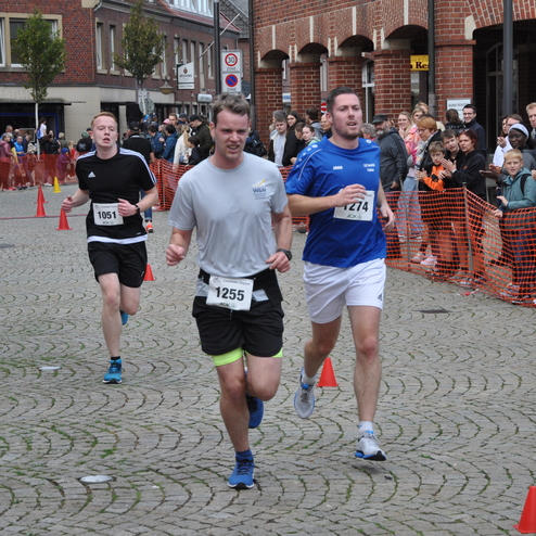 Drei L&auml;ufer in Sportkleidung sprinten auf einer gepflasterten Stra&szlig;e, umgeben von einer Menschenmenge und Absperrungen.