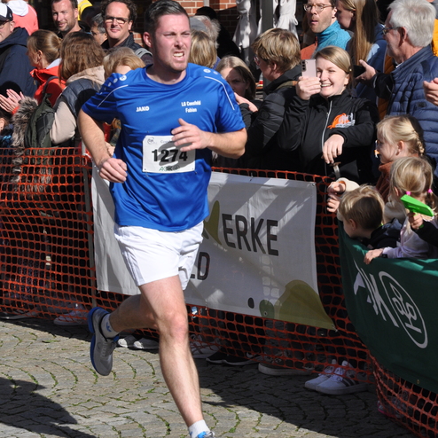 Mann in blauer Sportkleidung mit Startnummer 1274 l&auml;uft bei einem Wettkampf, umgeben von jubelnden Zuschauern.