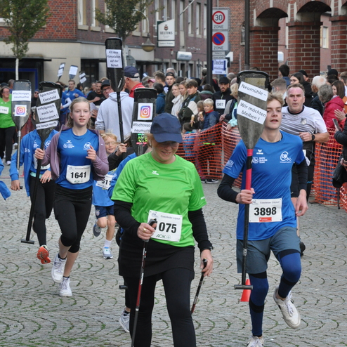 Vier L&auml;ufer mit Walkst&ouml;cken und Nummernshirts nehmen an einem Wettkampf in einer belebten Stra&szlig;e teil.