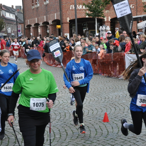 L&auml;uferinnen und L&auml;ufer mit Startnummern und Paddeln beim Wettlauf vor jubelndem Publikum auf einer Pflasterstra&szlig;e.