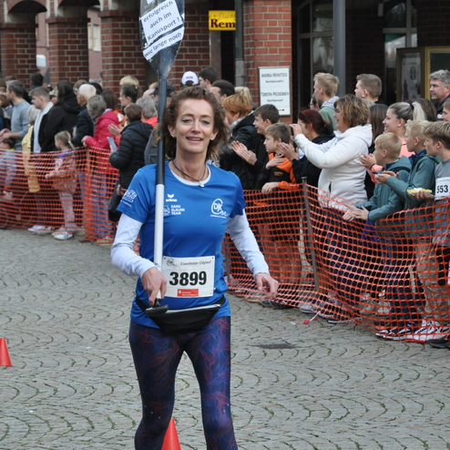 Eine L&auml;uferin in blauer Sportkleidung tr&auml;gt ein Schild und l&auml;uft durch eine jubelnde Menge bei einem Stra&szlig;enlauf.