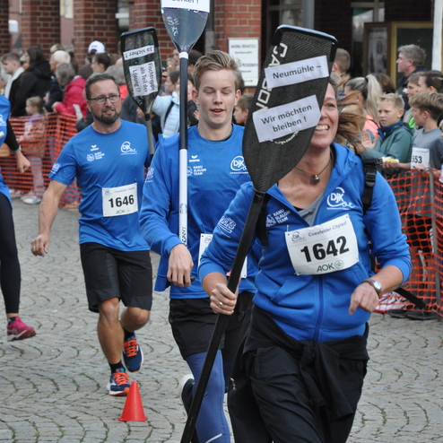 Teilnehmer eines Laufs tragen Kanusport-Paddel mit Plakaten, um einen Kanuslalom zu feiern, umringt von Zuschauern.