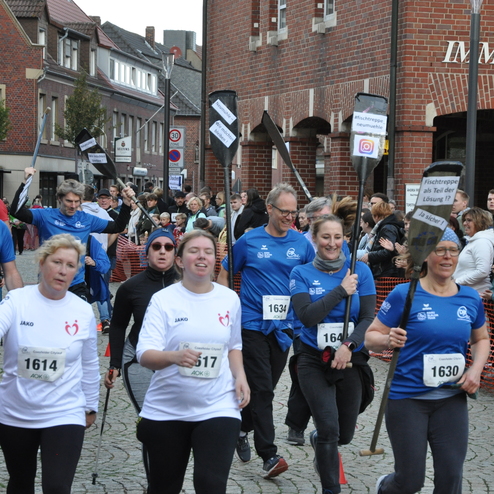 Gruppe von L&auml;ufern in blauen T-Shirts mit Paddeln, umgeben von Zuschauern auf einer Stra&szlig;enveranstaltung.