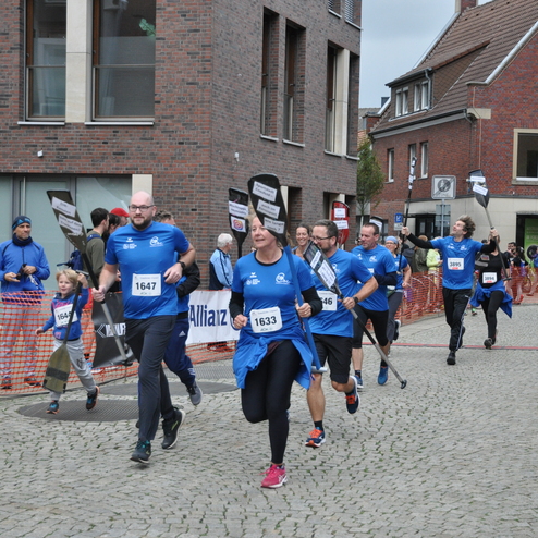 Gruppe von L&auml;ufern in blauen T-Shirts sprintet auf gepflasterter Stra&szlig;e w&auml;hrend eines Wettkampfs. Zuschauer applaudieren.