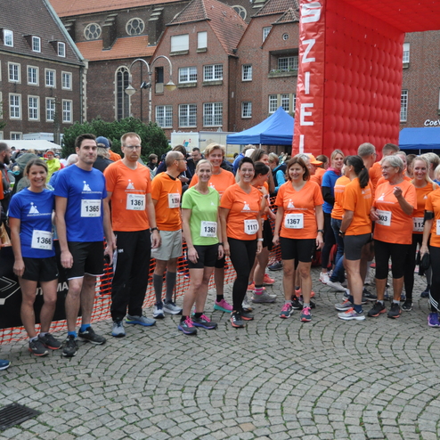 Gruppe von L&auml;ufern in bunter Sportkleidung steht am Startpunkt eines Wettlaufs, umgeben von Zuschauern und St&auml;nden.