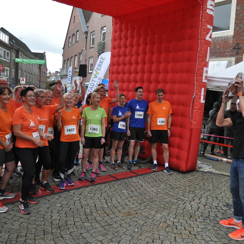 Gruppenshooting von L&auml;ufern in orange und blauen Shirts vor einem roten Startbogen bei einer Laufveranstaltung.