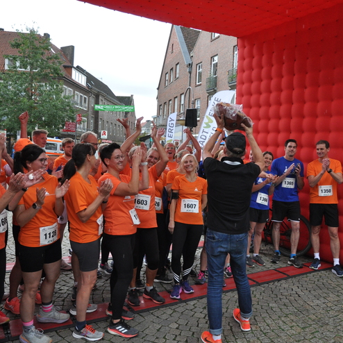 Gruppierung von L&auml;ufern in orange T-Shirts, die jubelnd eine Auszeichnung unter einem roten Zielbogen entgegennehmen.