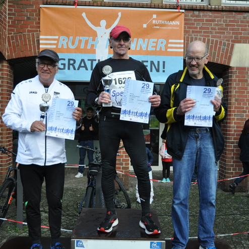 Drei M&auml;nner stehen auf dem Siegerpodest, halten Pokale und Urkunden nach einem Laufwettbewerb, im Hintergrund ein Veranstaltungsbanner.