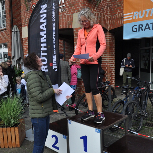 Frau in orangefarbener Jacke steht auf dem Siegerpodest und erh&auml;lt eine Urkunde von einer Person im Vordergrund.