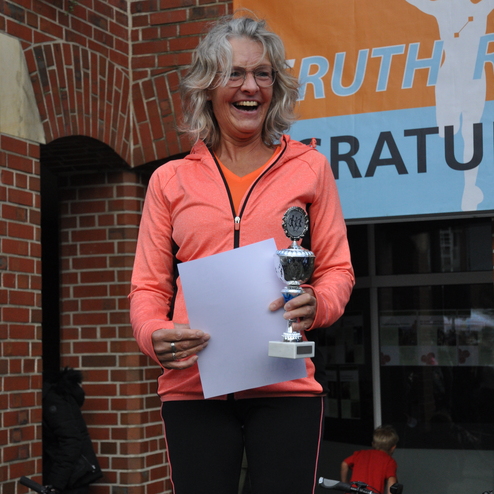 Frau in orangener Sportjacke l&auml;chelt, h&auml;lt eine Urkunde und einen Pokal auf einer Siegerehrung vor einer Backsteinwand.