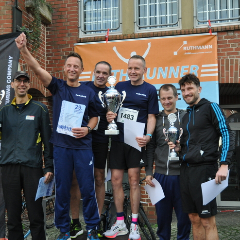 Sechs M&auml;nner in Sportkleidung stehen mit Pokalen und Urkunden vor einem Veranstaltungsbanner nach einem Lauf.