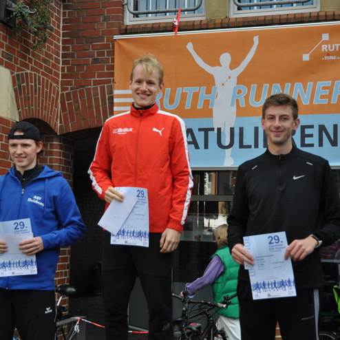 Drei L&auml;ufer stehen auf dem Siegerpodest, halten Urkunden und l&auml;cheln vor einem Banner mit der Aufschrift &bdquo;Ruth Runner&ldquo;.