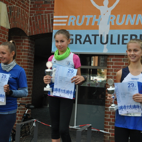 Drei junge L&auml;uferinnen stehen mit Pokalen und Urkunden vor einer Veranstaltungstafel. Siegerinnen des 29. Coesfelder Stadtlaufs.