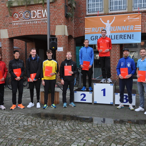 Gruppe von Sportlern steht auf dem Podium und hält Urkunden nach einem Laufwettbewerb vor einem Gebäude.