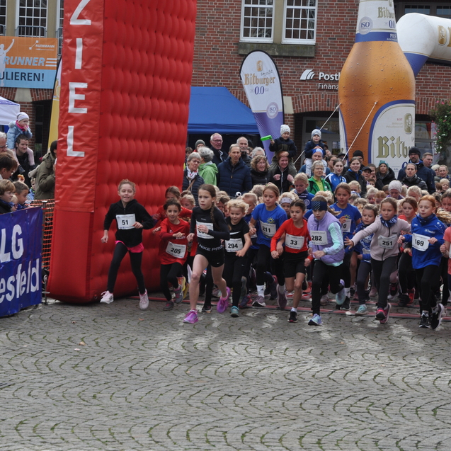 Start eines Kinderlaufs mit vielen laufenden Mädchen vor einem großen Zielbogen in einer belebten Stadt.
