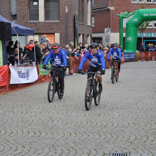 Gruppe von Mountainbikern in blauen Trikots fährt in eine Menschenmenge vor einem Zielbogen bei einer Fahrradveranstaltung.