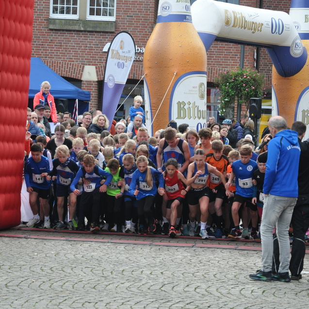 Gruppierung von jugendlichen Läufern am Start einer Veranstaltung, umgeben von Zuschauern und einem Zielbogen.