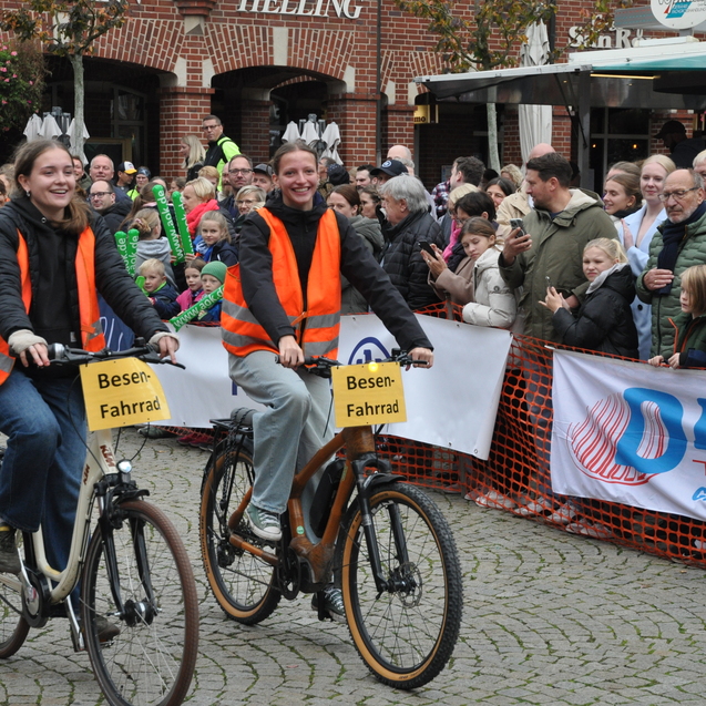 Zwei Radfahrer in orangefarbenen Westen fahren auf Besen-Fahrrädern vor einer jubelnden Menschenmenge.