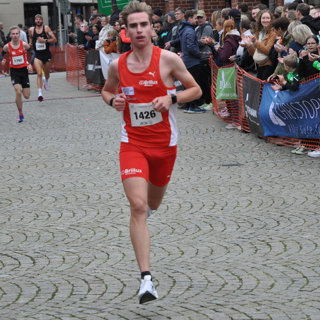 Junger Läufer in rotem Trikot mit Nummer 1426 überquert das Ziel bei einem Lauf, Zuschauer applaudieren.