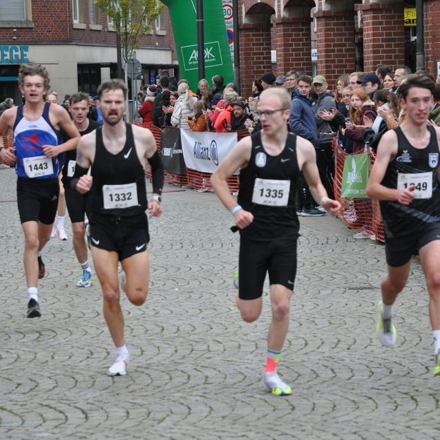 Vier Läufer sprinten über eine gepflasterte Straße während eines Wettkampfs, umgeben von jubelndem Publikum.