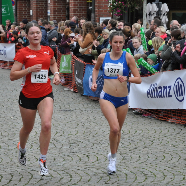 Zwei weibliche L&auml;uferinnen im Wettkampf, eine in rotem Shirt, die andere in blauem, umgeben von jubelnden Zuschauern.