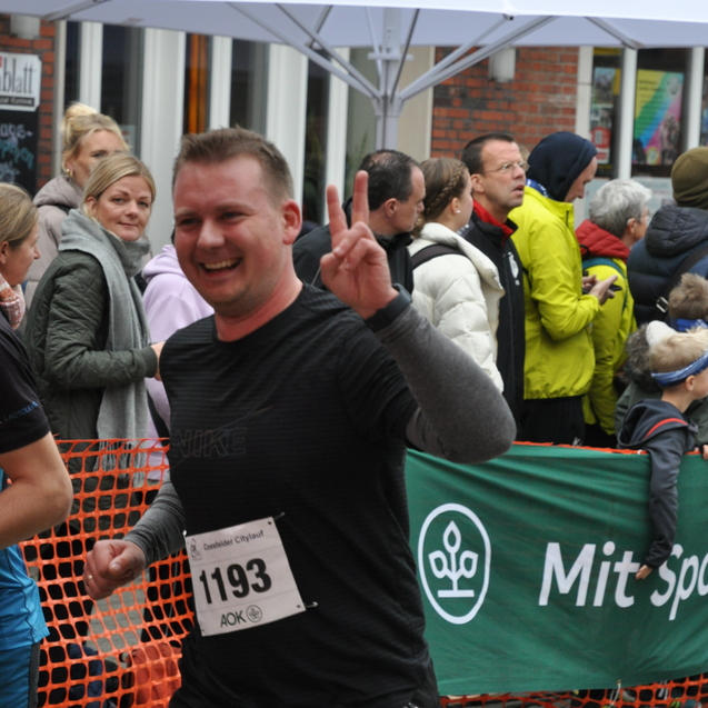 L&auml;ufer in schwarzem Shirt mit Startnummer 1193 zeigt ein Victory-Zeichen w&auml;hrend des Stadtlaufs, im Hintergrund Menschenmenge.