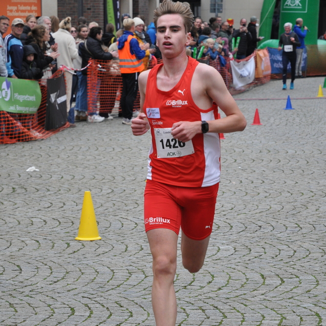 Junger L&auml;ufer in rotem Sportanzug sprintet auf einem Pflasterweg, umgeben von zuschauenden Menschen und Absperrungen.