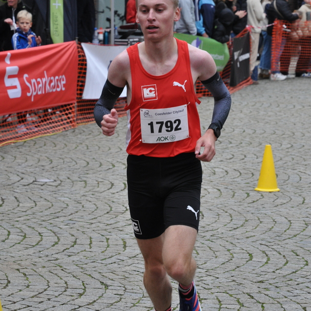 Junger L&auml;ufer in rotem Trikot mit Startnummer 1792 sprintet auf einer Pflasterstra&szlig;e, Zuschauer im Hintergrund.