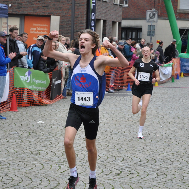 Junger Athlet jubelt beim Zieleinlauf eines Wettlaufs, w&auml;hrend eine weitere L&auml;uferin im Hintergrund vorbei l&auml;uft.