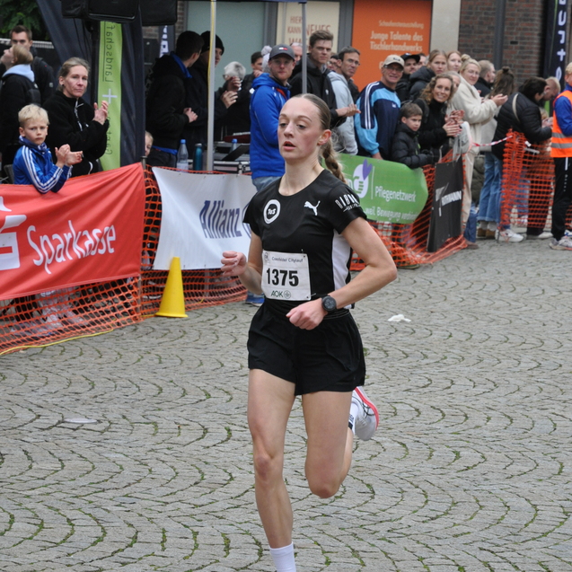 L&auml;uferin in schwarzer Sportkleidung mit Startnummer 1375 sprintet auf einem gepflasterten Weg, umgeben von Zuschauern.