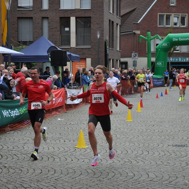 Zwei L&auml;ufer erreichen das Ziel eines Stra&szlig;enrennens, umgeben von Zuschauern und einem gr&uuml;nen Zielbogen.