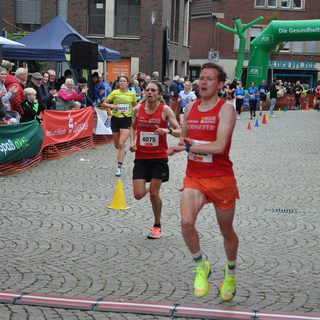 Drei L&auml;ufer &uuml;berqueren die Ziellinie eines Stadtmarathons, umgeben von Zuschauern und bunten Banner-Displays.