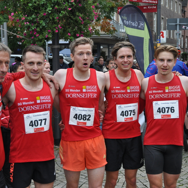 F&uuml;nf L&auml;ufer in roten Trikots mit Startnummern stehen l&auml;chelnd auf einer Veranstaltung, im Hintergrund Zuschauer und Stadtansicht.