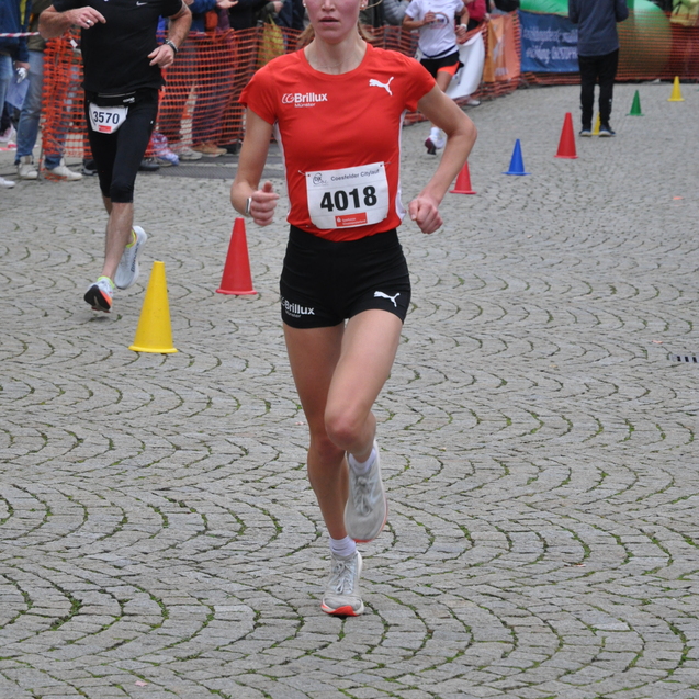 Junge Frau in rotem Shirt l&auml;uft bei einem Stra&szlig;enlauf, im Hintergrund Zuschauer und andere L&auml;ufer.