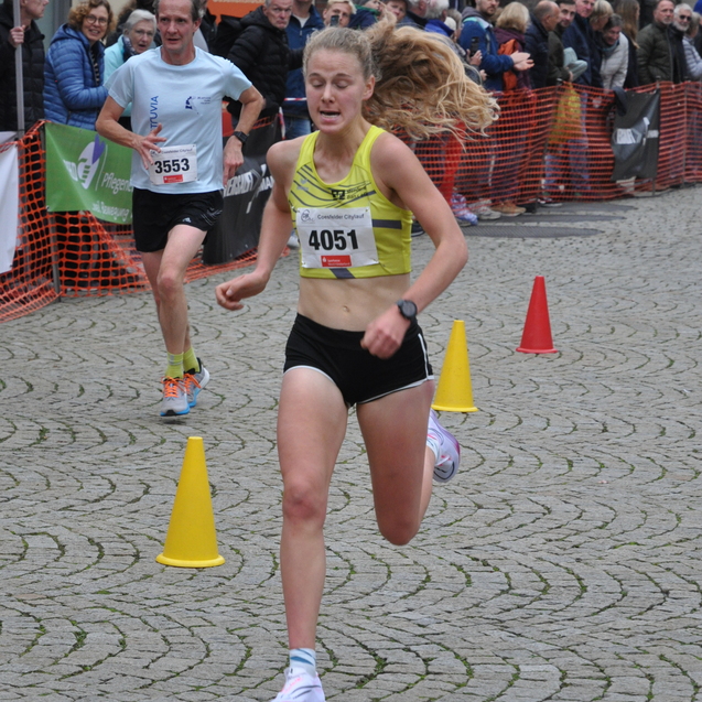 L&auml;uferin in gelber Sportkleidung sprintet auf Pflastersteinstra&szlig;e, Zuschauer sind im Hintergrund sichtbar.