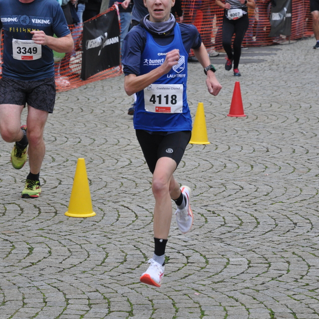 L&auml;ufer in blauer Sportkleidung sprintet bei einem Wettlauf auf gepflastertem Boden mit Absperrungen und Zuschauern im Hintergrund.