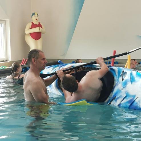 Zwei Männer stehen in einem Schwimmbecken, einer hilft dem anderen beim Einsteigen in ein Kanu. Im Hintergrund sind Kinder.