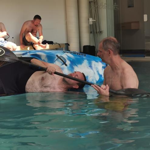 Zwei Männer im Wasser, einer liegt im Kajak und erhält Unterstützung vom anderen, während ein weiterer Mann zuschaut.