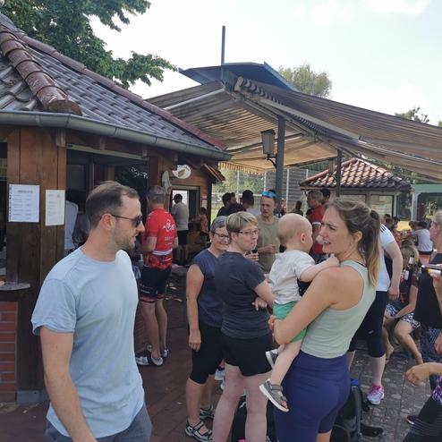 Menschen stehen in Gruppen vor einem Kiosk, einige trinken Bier, während andere mit Kindern sprechen.