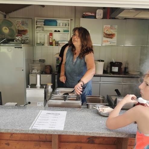 Frau in einer Imbissbude serviert Speisen, während ein Kind neben ihr auf Ketchup wartet. Preisliste auf der Theke sichtbar.