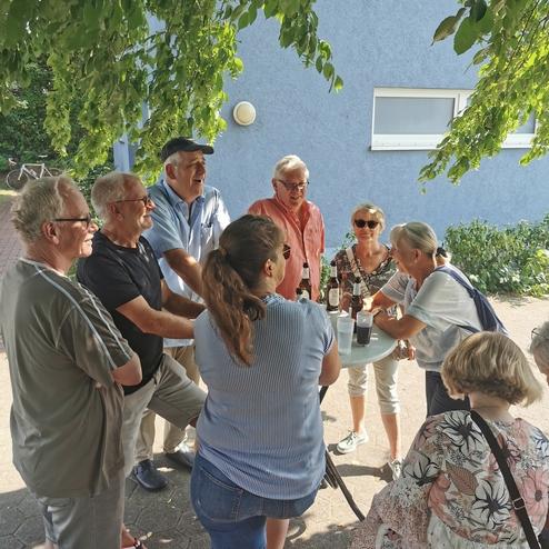 Gruppe von acht Personen bei einem geselligen Treffen unter einem Baum, mit Flaschen und Gläsern auf einem Tisch.